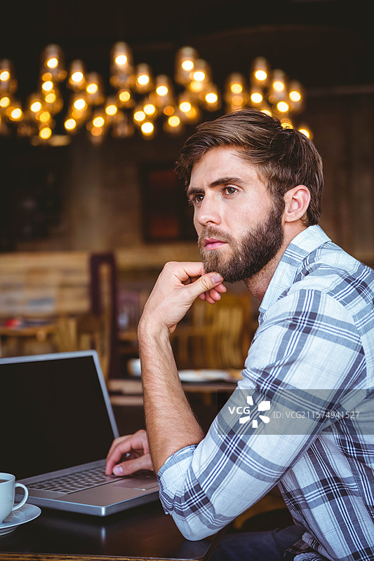 在咖啡馆的木桌上，男人一边喝咖啡一边使用笔记本电脑，周围有吊灯，留有空白空间。图片素材