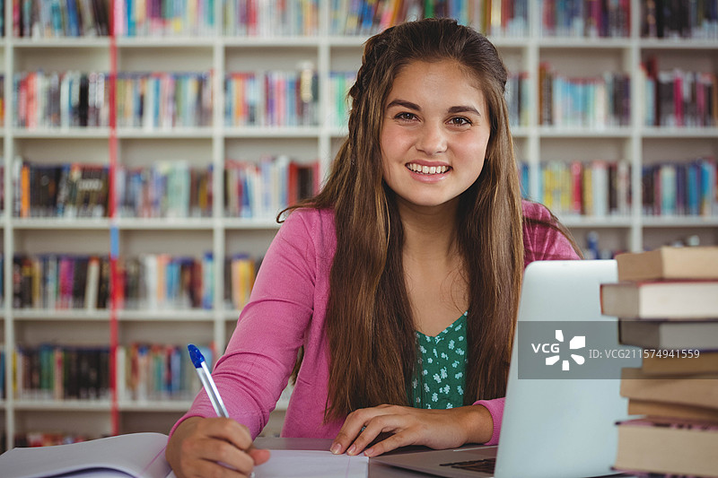 在图书馆的桌子上，青少年女孩正在写笔记，周围有书架、笔记本、钢笔、笔记本电脑和教科书。图片素材