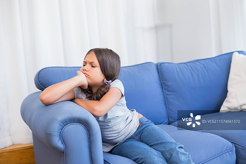 女孩坐在家里的蓝色沙发上，靠着手肘，侧目凝视，旁边有白色的枕头，留有空白空间。图片素材