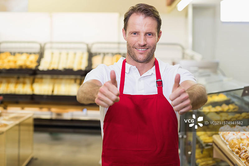 成熟的成年男子穿着红色围裙，在面包店柜台后面竖起大拇指，周围是面包和糕点。图片素材