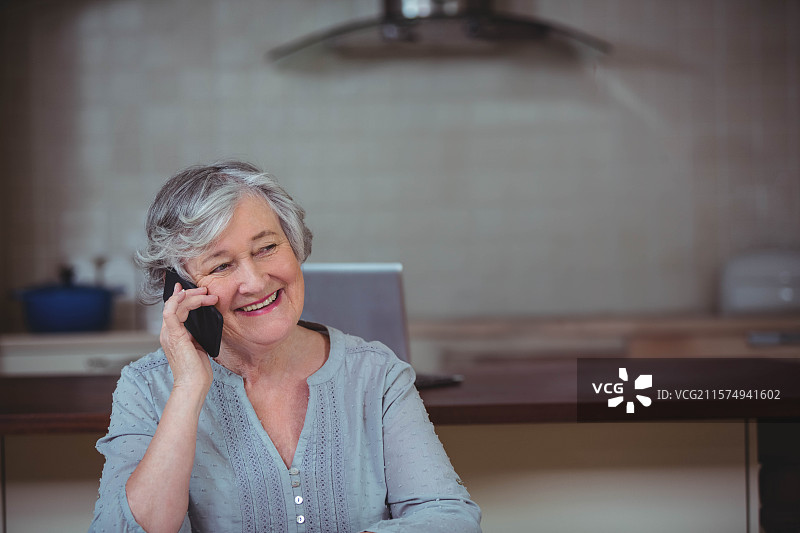 一位老年女性坐在厨房岛上，正在用智能手机通话，旁边有笔记本电脑、锅和抽油烟机。图片素材