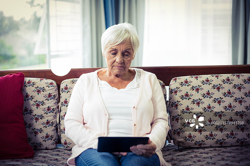 一位年长女性坐在花卉沙发上，手持黑色平板电脑，旁边是轻薄的窗帘。图片素材