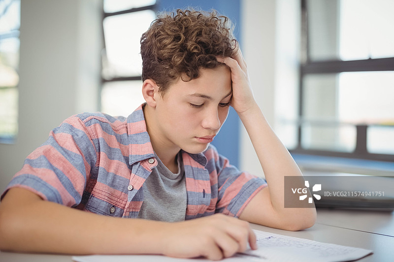 穿着条纹衬衫的少年向前倾身，在学校桌子上用笔记本写字。图片素材