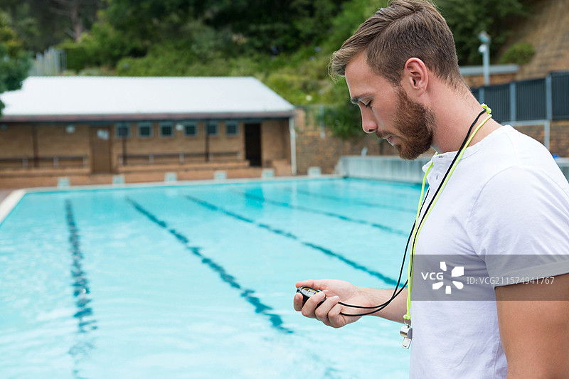 男教练手持秒表，查看显示屏，站在泳池边，旁边有泳道绳，留有空白空间。图片素材