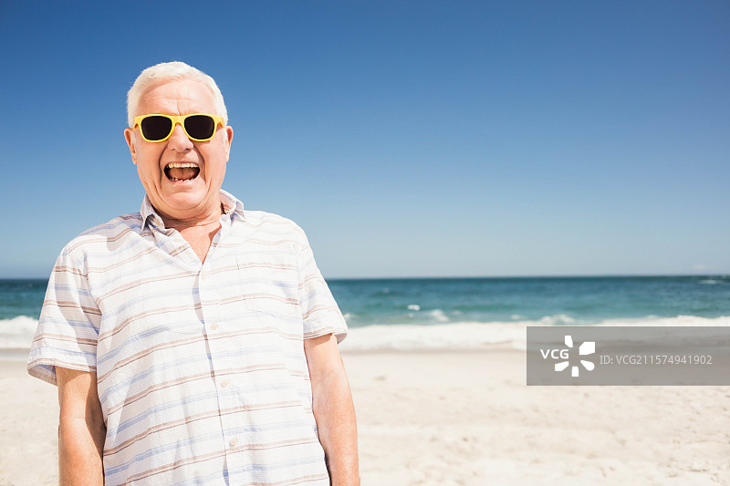 穿着条纹衬衫和黄色太阳镜的老年男子站在沙滩的海岸上，留有空白空间。图片素材