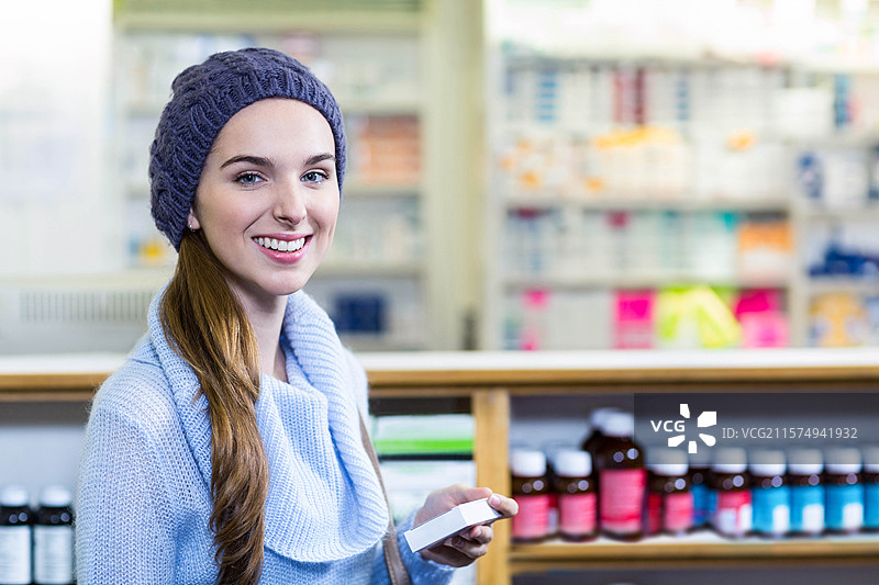 穿着毛线帽和毛衣的少女在药店柜台前微笑着手持药品盒。图片素材