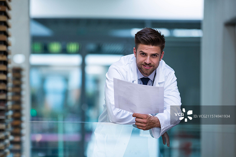穿着白色实验室外套的成年男子靠在设施走廊的玻璃栏杆上，手里拿着文件。图片素材