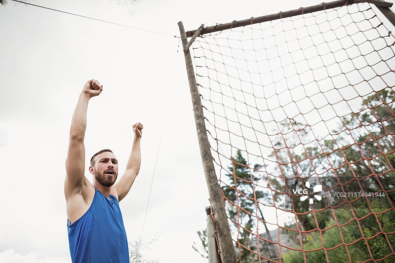 男子在树林空地旁的绳网障碍物旁举起双臂，周围有木柱。图片素材
