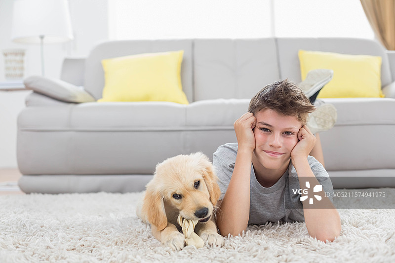 男孩躺在客厅的地毯上，旁边是灰色沙发，小狗正在啃咬绳结玩具。图片素材