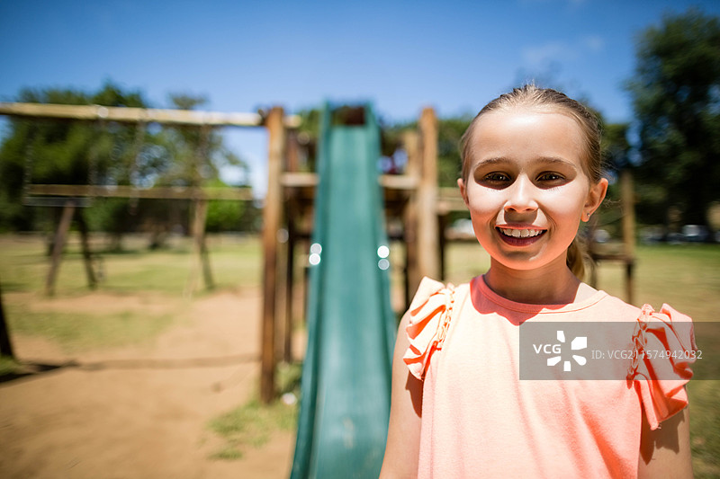 在公共公园里，站在木制游乐设施前的小女孩，旁边有绿色滑梯，留有空白空间。图片素材