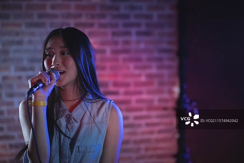 亚洲女性在俱乐部舞台上穿着无袖牛仔上衣，站在麦克风前唱歌，留有空白空间。图片素材