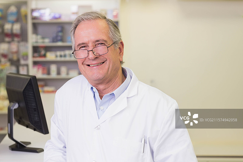 穿着实验室外套的老年男性药剂师站在药房的柜台后，使用电脑显示器。图片素材