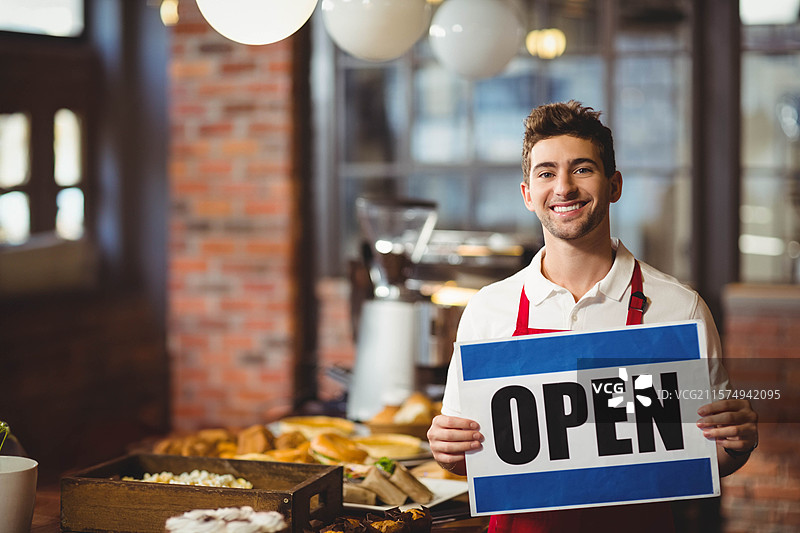 男咖啡师站在面包店柜台后，手持“开放”标志，正在摆放点心托盘。图片素材