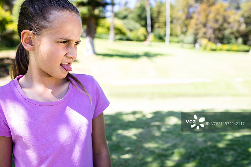 穿着紫色T恤的女孩在阳光明媚的公园里做鬼脸，吐出舌头，向左看，留有空白空间。图片素材