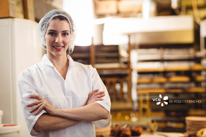 女糕点师穿着厨师外套和发网，站在面包店的货架和托盘之间，留有空白空间。图片素材