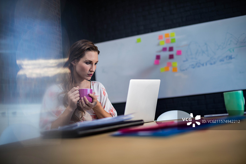 女性在现代办公室的木桌上，手握紫色杯子，正在笔记本电脑上打字，桌上有便利贴。图片素材