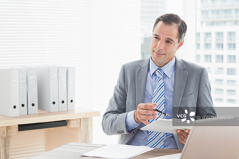 穿着灰色西装、条纹领带的男子手持夹板在办公桌前审阅文件，留白空间图片素材