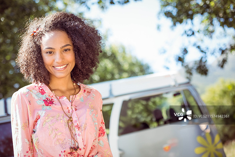 穿着粉色衬衫的非裔美国女性站在露营地的复古房车旁，房车上贴有花朵贴纸。图片素材