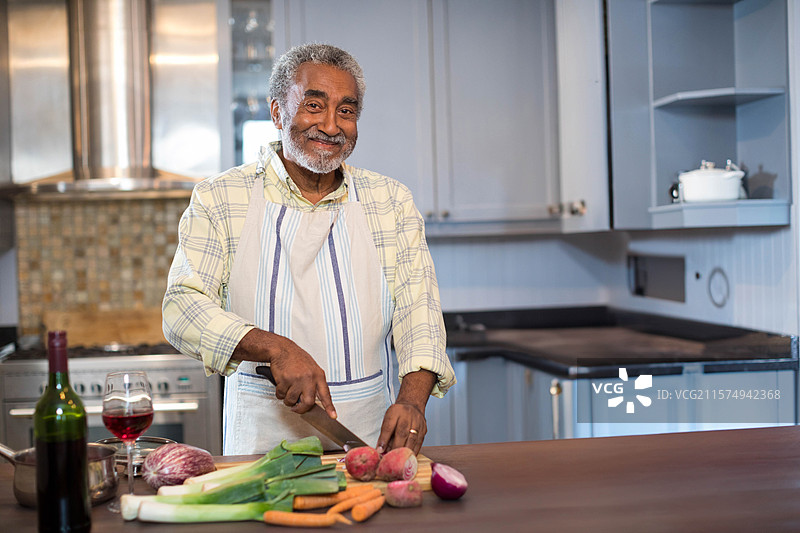 一位年长的非裔美国男性在厨房的木砧板上切菜，旁边有一瓶葡萄酒。图片素材