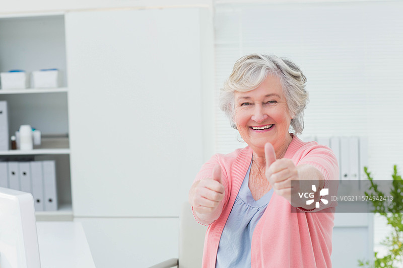 一位年长女性坐在白色桌子旁，双臂举起，做出竖起大拇指的手势，面前有一台显示器。图片素材
