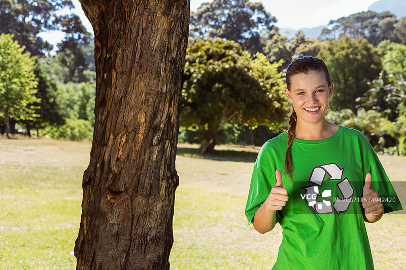 穿着绿色回收衫的女人站在阳光明媚的草地上，旁边是一棵树干，留有空白空间。图片素材