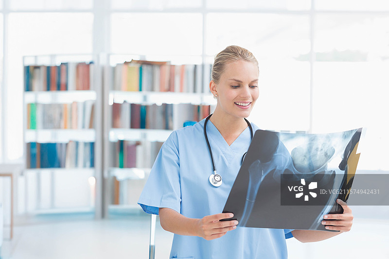 穿着蓝色护士服的女性佩戴听诊器，手持盆腔X光片，在书架旁的阅片室内。图片素材