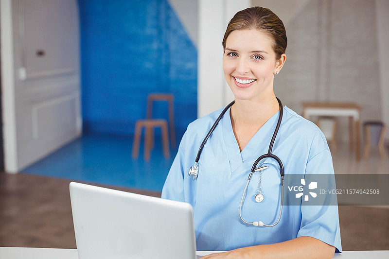 穿着护士服和听诊器的女性护士坐在接待处的桌子前使用笔记本电脑，留有空白空间图片素材