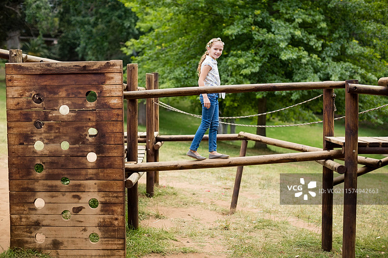 女孩子在公园游乐场的障碍课程上沿着木梁平衡，旁边有绳索护栏。图片素材