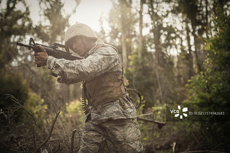 穿着战术背心和头盔的士兵，在茂密的林地中移动，手持步枪向前侦查。图片素材