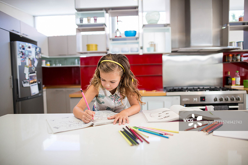 女孩坐在厨房的白色台面岛上，用彩色铅笔给工作簿上色。图片素材