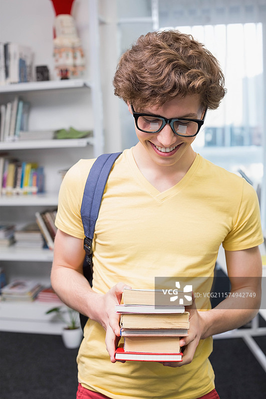 男学生微笑着站在图书馆里，手里拿着五本书，背着书包。图片素材