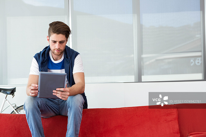 男子坐在明亮的休息区红色沙发上使用平板电脑，旁边有一个自行车框架。图片素材