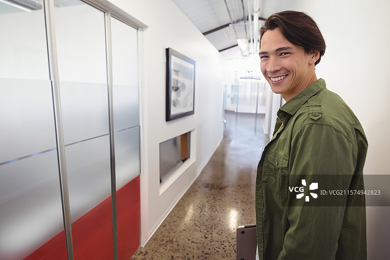 亚洲男子微笑着手持合上的笔记本电脑，站在现代办公室走廊中，留有空白空间。图片素材