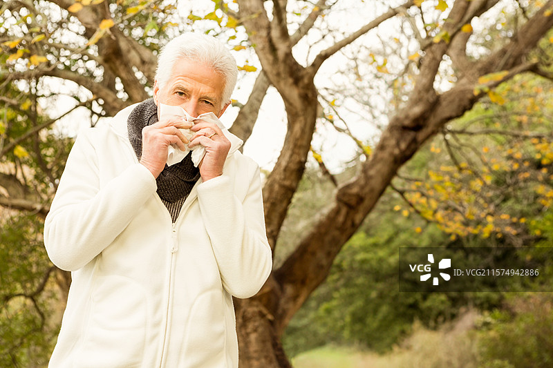 秋天，一位穿着白色夹克的老人在公园里站着，手里拿着纸巾捂住鼻子，留有空白空间。图片素材