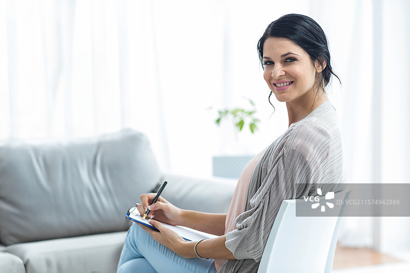 女人坐在白色椅子上，侧身用笔在夹板上写笔记，身处明亮的家庭办公室。图片素材