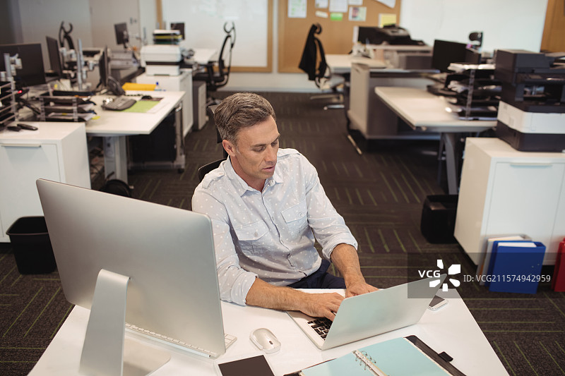 中年男士穿着商务休闲服装，在开放式办公室的桌子上使用笔记本电脑，旁边有显示器。图片素材