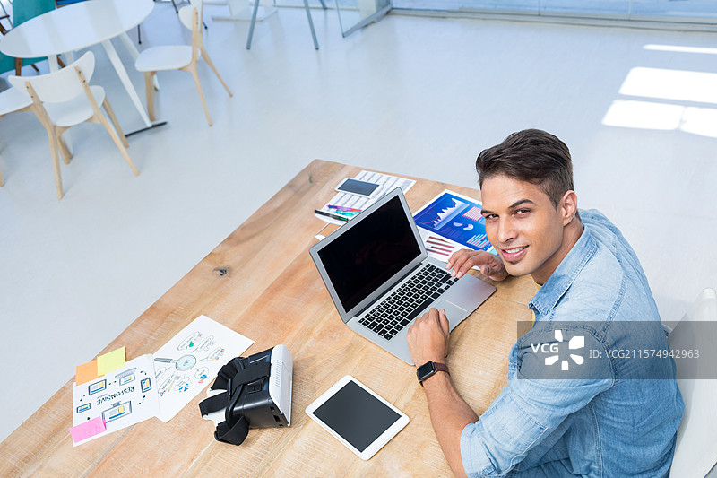 中年男子穿着牛仔衬衫，在办公室里佩戴虚拟现实头盔使用笔记本电脑，留有空白空间。图片素材