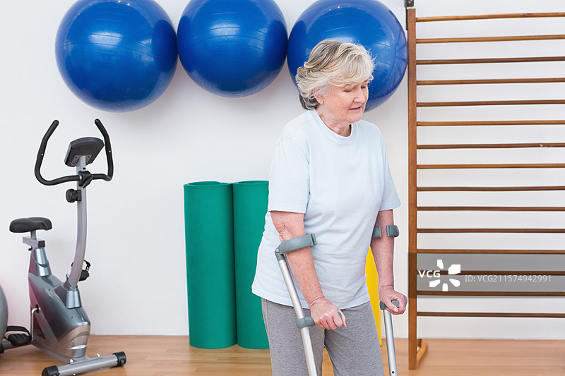 一位年长的白人女性在康复健身房中使用前臂拐杖保持平衡，周围有球和自行车，留有空白空间。图片素材
