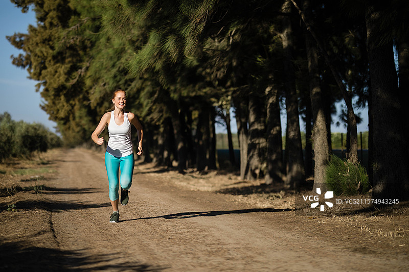 穿着白色无袖上衣和青绿色紧身裤的女性跑者在森林边缘的泥土小径上慢跑图片素材