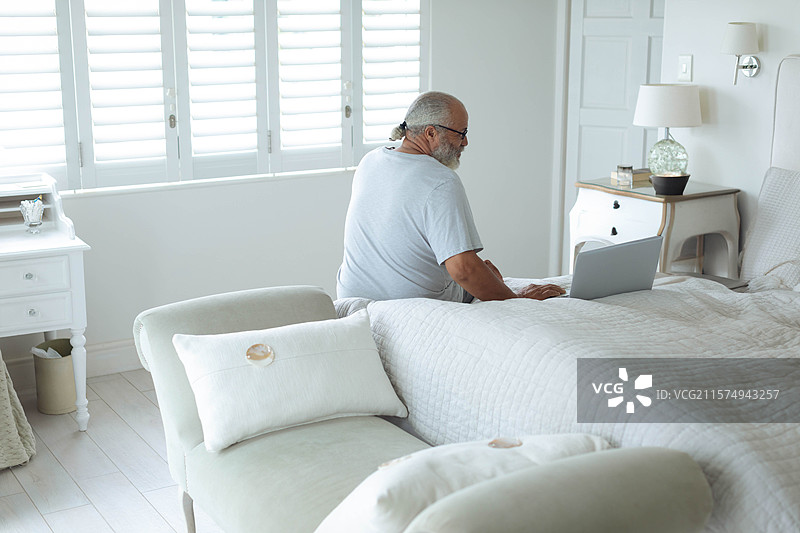 老年男子在简约风格的卧室里，床上用笔记本电脑工作，房间有白色百叶窗和床头柜。图片素材