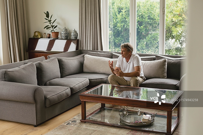 老年男子坐在客厅的沙发上，手势指向玻璃咖啡桌，留有空白空间图片素材