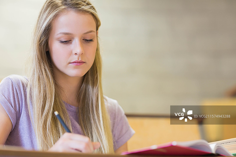 穿着淡紫色T恤的少女在教室桌子上用铅笔写笔记，旁边留有空白空间。图片素材