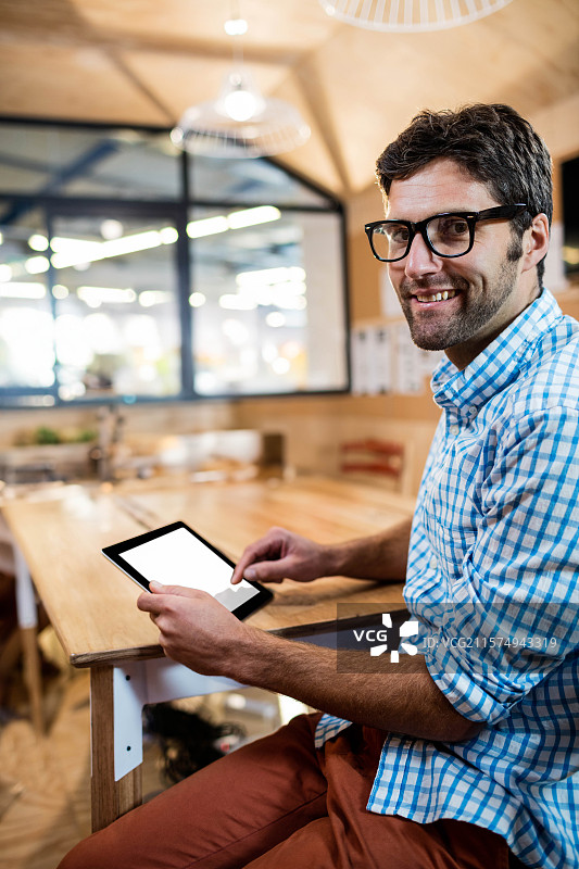 男子在现代共享办公室的玻璃隔断旁的木桌上使用平板电脑，留白空间。图片素材