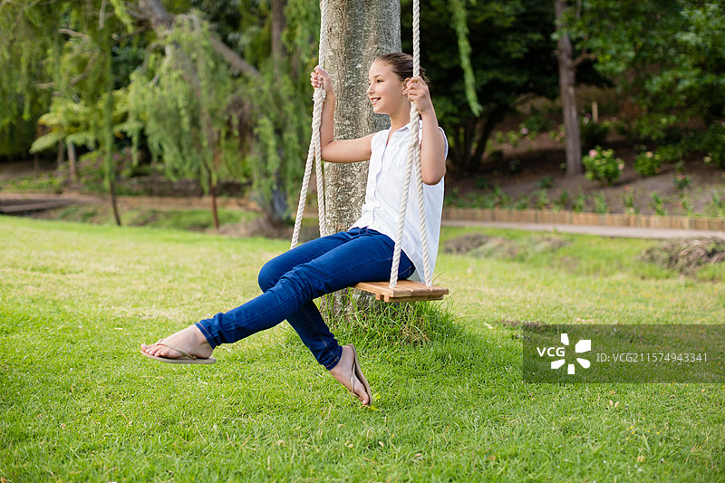 小女孩在后院的木板秋千上荡秋千，粗绳子和结实的树干支撑着她。图片素材
