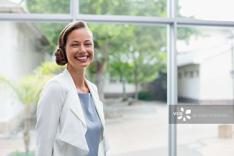 二十多岁的女性微笑着站在大堂的玻璃面板旁，穿着干净利落的白色西装外套。图片素材