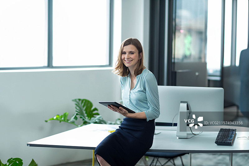 女人坐在白色桌子的边缘，手里拿着平板电脑，旁边有显示器、键盘和盆栽植物，身处办公室。图片素材