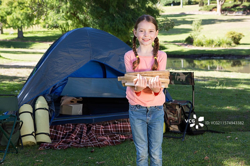 小女孩手握柴火束，站在草坪上的蓝色圆顶帐篷前。图片素材