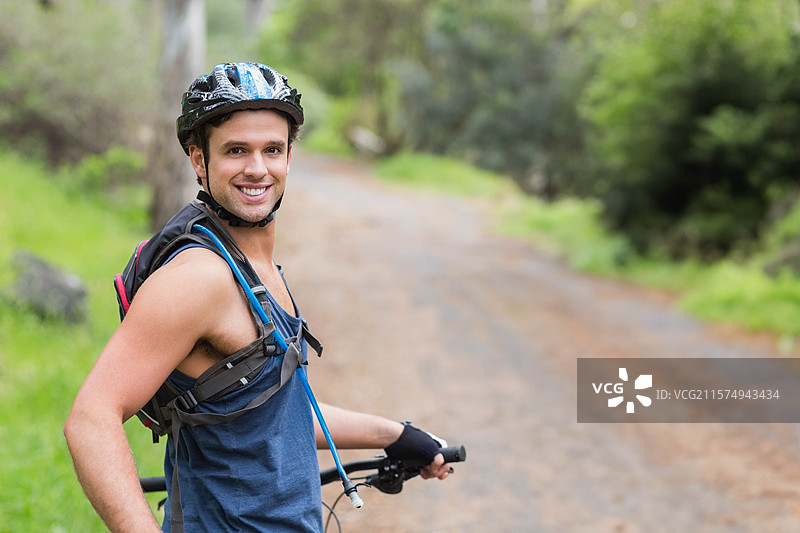 男子微笑着握住自行车把手，穿着头盔和水袋背包，站在森林小径上，留有空白空间。图片素材