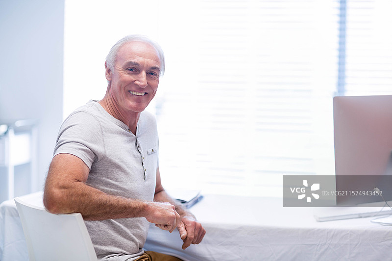 微笑的老年男子在家中的办公室里使用桌面电脑，旁边有智能手机和窗帘。图片素材