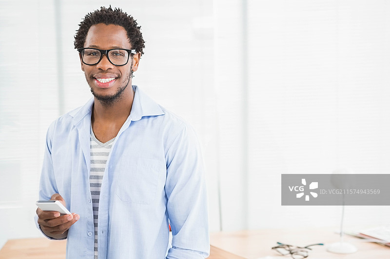 一位非裔美国男性站在木桌旁，手持智能手机，周围有文件和台灯，身处办公室。图片素材